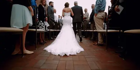 A bride in a white lace gown walking down a brick aisle toward a wedding ceremony with guests seated on either side.