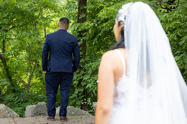 A bride and groom standing outdoors in a wooded area, with the groom facing away and the bride in the foreground.
