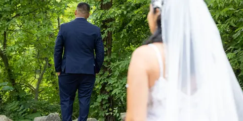A bride and groom standing outdoors in a wooded area, with the groom facing away and the bride in the foreground.