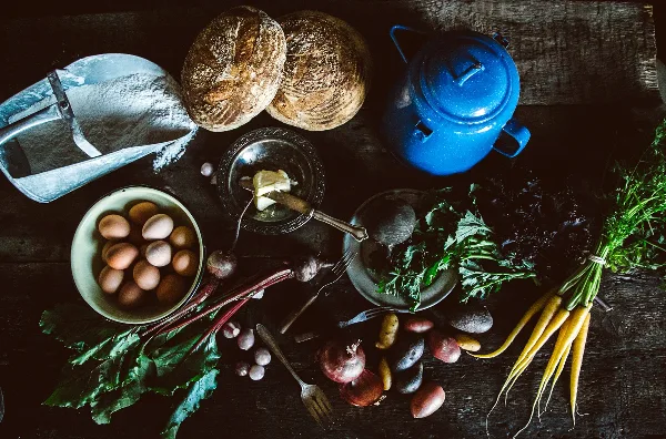 Rustic table setting with loaves of bread, butter, eggs, and fresh vegetables and herbs.
