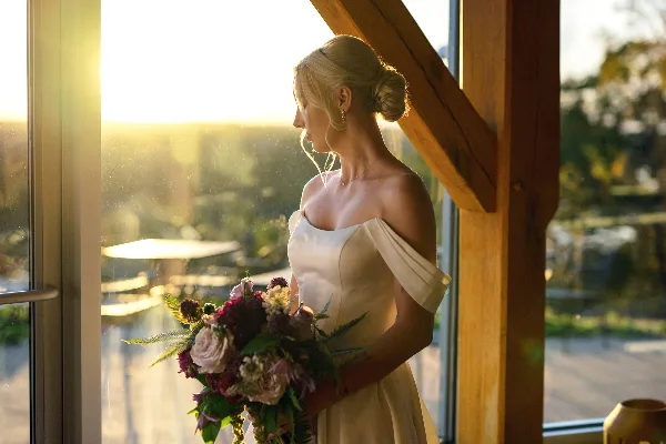 A bride in a white off-the-shoulder gown stands by a window at sunset, holding a floral bouquet.