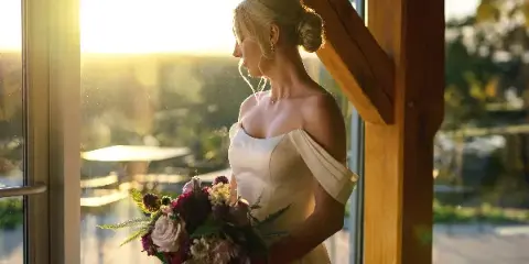 A bride in a white off-the-shoulder gown stands by a window at sunset, holding a floral bouquet.