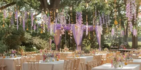 Outdoor wedding reception under oak trees featuring purple floral drapery, string lights, and white tablecloths.