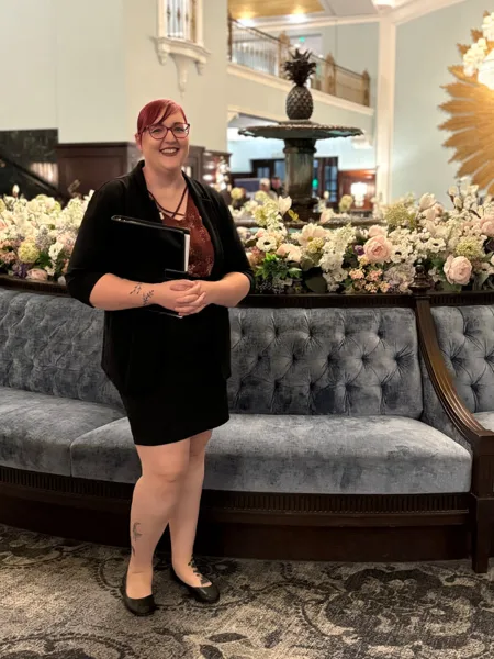 A woman with red hair and glasses stands in a formal venue with floral arrangements, holding a black binder.