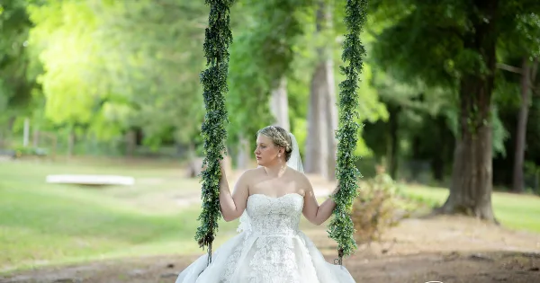 A bride in a white lace wedding dress sitting on a swing decorated with greenery in a wooded outdoor area.