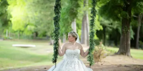 A bride in a white lace wedding dress sitting on a swing decorated with greenery in a wooded outdoor area.