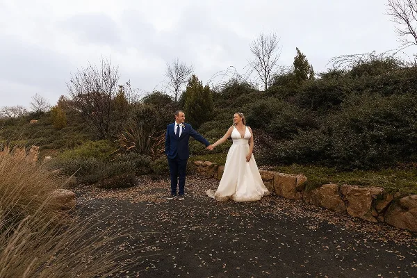 A bride and groom holding hands in a natural outdoor setting with a stone wall and vegetation.