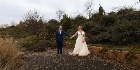 A bride and groom holding hands in a natural outdoor setting with a stone wall and vegetation.