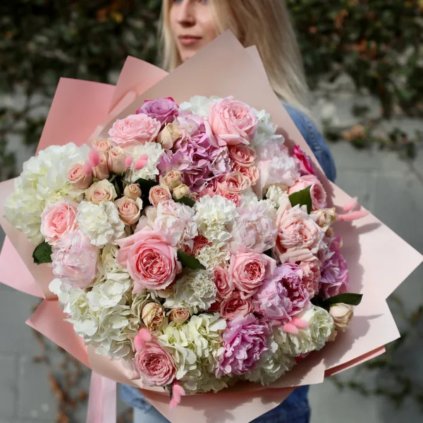 A large, hand-tied bouquet of pink garden roses, white hydrangeas, and lisianthus wrapped in pink paper.