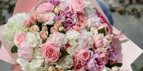 A large, hand-tied bouquet of pink garden roses, white hydrangeas, and lisianthus wrapped in pink paper.