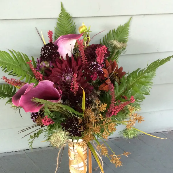 A vibrant floral arrangement with pink calla lilies, dark red dahlias, and green ferns in a gold-wrapped vase.