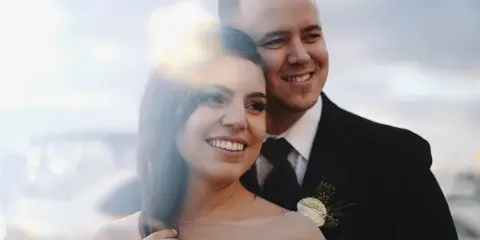 A cinematic close-up of a smiling bride and groom in wedding attire, with a soft lens flare and blurred background.