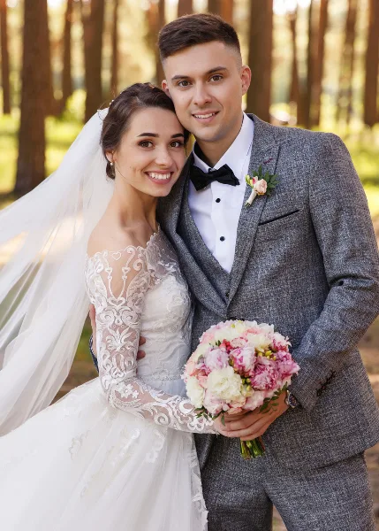 A smiling bride and groom in wedding attire standing together outdoors holding a bouquet.