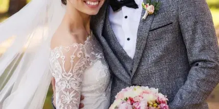 A smiling bride and groom in wedding attire standing together outdoors holding a bouquet.