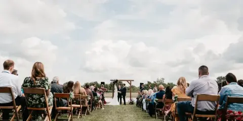 Outdoor wedding ceremony with a bride and groom at an altar and seated guests in a grassy field.