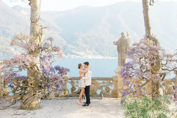 A bride and groom standing together on a terrace overlooking a lake and mountains, framed by blooming wisteria trees.