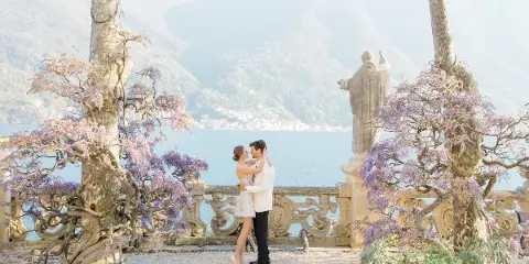 A bride and groom standing together on a terrace overlooking a lake and mountains, framed by blooming wisteria trees.