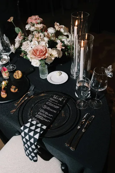 A professionally styled wedding table setting featuring a black tablecloth, floral centerpiece, tall candles, and a custom menu card.