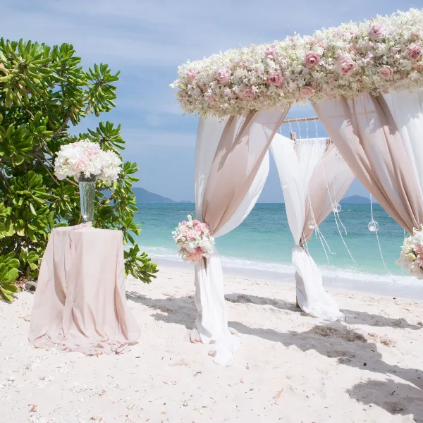 A wedding floral arch with white and pink flowers and draped fabric set on a beach, next to a pedestal with a floral arrangement.