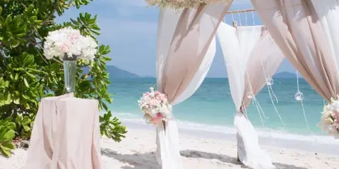 A wedding floral arch with white and pink flowers and draped fabric set on a beach, next to a pedestal with a floral arrangement.