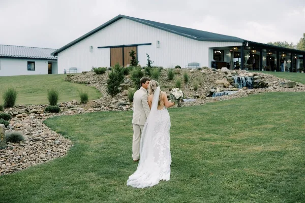 A bride and groom standing on a lawn in front of a modern white wedding venue building with a stone water feature.