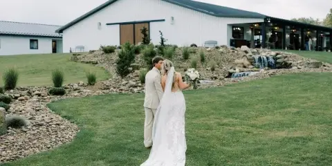 A bride and groom standing on a lawn in front of a modern white wedding venue building with a stone water feature.