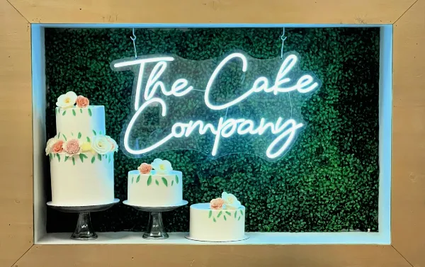 A bakery storefront window displaying a neon sign reading 'The Cake Company' and three white cakes decorated with floral accents.