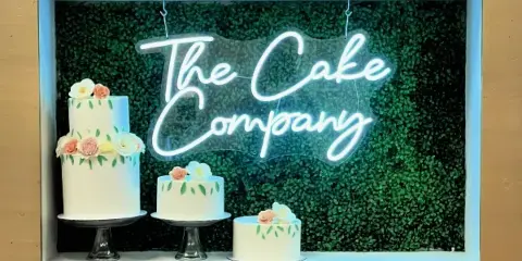 A bakery storefront window displaying a neon sign reading 'The Cake Company' and three white cakes decorated with floral accents.