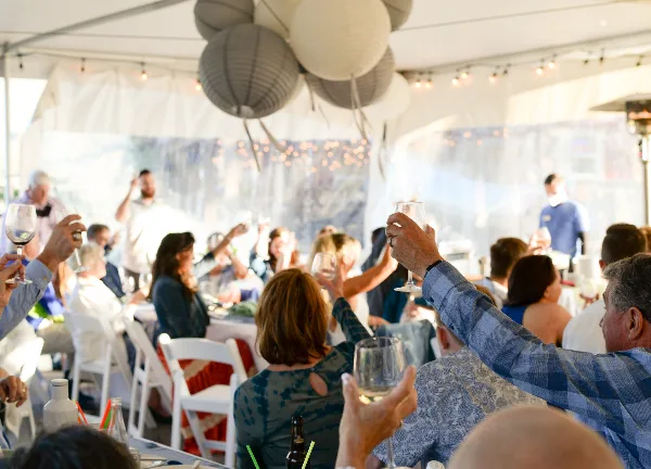 Guests at a wedding reception raising wine glasses in a toast under a white tent.