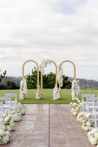 Outdoor wedding ceremony setup featuring a floral arch, white chairs, and aisle flowers on a paved area.
