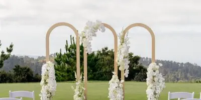 Outdoor wedding ceremony setup featuring a floral arch, white chairs, and aisle flowers on a paved area.