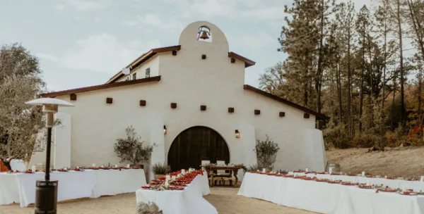 A white stucco building with an arched entryway set up for a wedding reception with long tables and floral centerpieces.