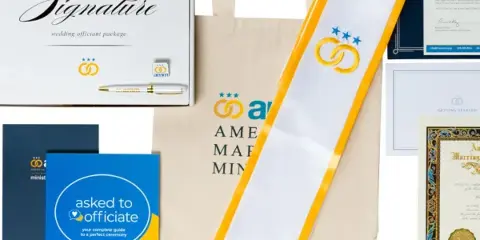 A flat lay of the American Marriage Ministries Signature Wedding Officiant Package, featuring a white and gold stole, an 'Asked to Officiate' workbook, a certificate, and a tote bag.