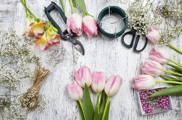 A flat lay of fresh pink and white tulips, orchids, and baby's breath arranged with floral design tools including shears, wire, and pins on a white wooden surface.