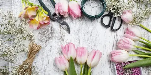 A flat lay of fresh pink and white tulips, orchids, and baby's breath arranged with floral design tools including shears, wire, and pins on a white wooden surface.