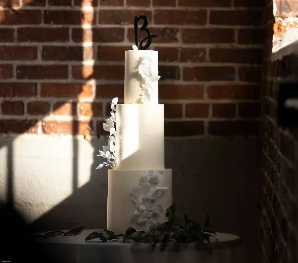A three-tier white wedding cake decorated with white flowers and a black monogram topper, set against a brick wall.