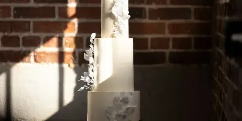 A three-tier white wedding cake decorated with white flowers and a black monogram topper, set against a brick wall.
