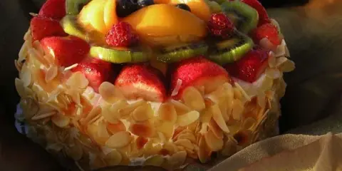 A round cake decorated with sliced strawberries, kiwi, blueberries, and toasted almonds, resting on a gold cloth.
