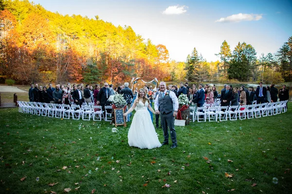 A bride and groom walking down the aisle during an outdoor autumn wedding ceremony with guests seated in white chairs.