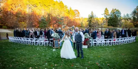 A bride and groom walking down the aisle during an outdoor autumn wedding ceremony with guests seated in white chairs.