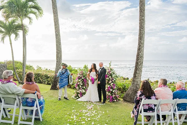 A bride and groom standing at an altar with an officiant and seated guests during a wedding ceremony on a grassy lawn overlooking the ocean.