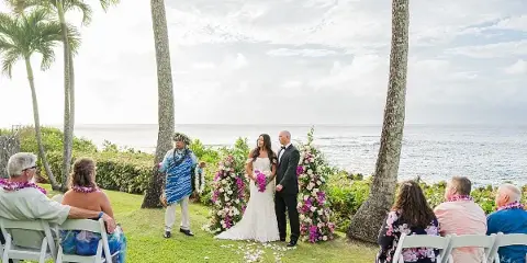 A bride and groom standing at an altar with an officiant and seated guests during a wedding ceremony on a grassy lawn overlooking the ocean.