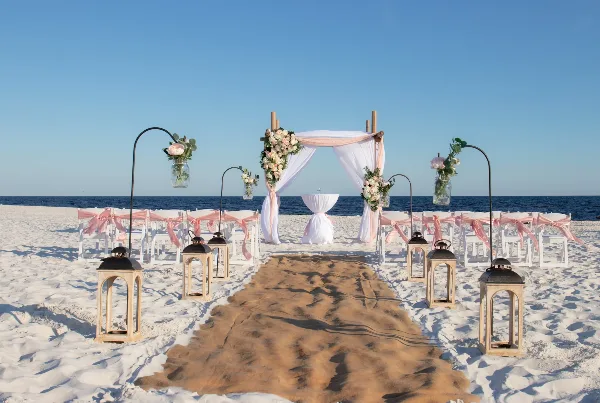 A beach wedding ceremony setup featuring a floral arch, white chairs with pink sashes, and a sandy aisle leading to the ocean.
