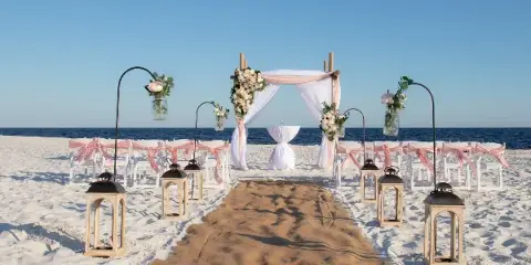 A beach wedding ceremony setup featuring a floral arch, white chairs with pink sashes, and a sandy aisle leading to the ocean.