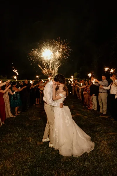 A bride and groom kissing during a sparkler exit at a wedding on a grassy field at night.