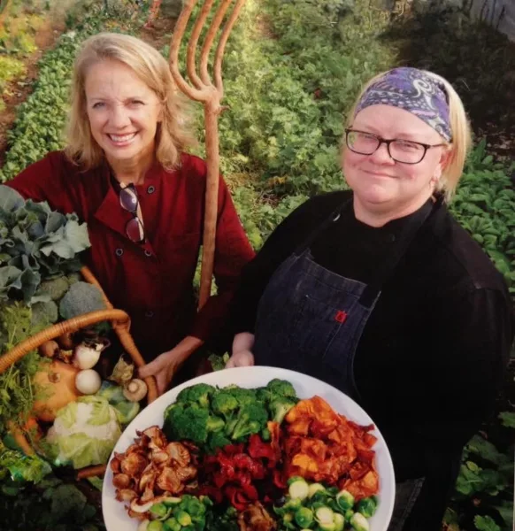 Two women standing in a garden holding a basket of fresh vegetables and a plate of prepared food.