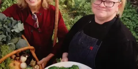 Two women standing in a garden holding a basket of fresh vegetables and a plate of prepared food.