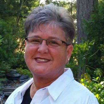 Portrait of a smiling woman with short gray hair and glasses wearing a white collared shirt, photographed outdoors in front of greenery.