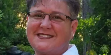 Portrait of a smiling woman with short gray hair and glasses wearing a white collared shirt, photographed outdoors in front of greenery.
