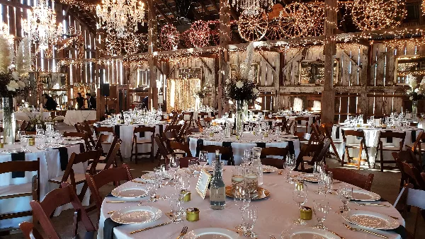 A rustic event venue interior featuring multiple round tables set with white linens, glassware, and centerpieces, surrounded by wooden chairs and string lights.